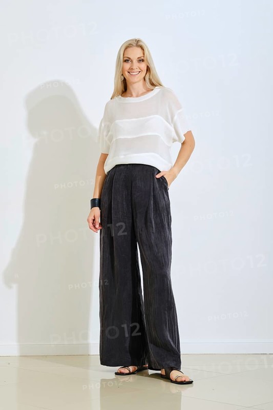 A model stands confidently indoors wearing a light top and loose black pants. The setting features soft lighting and a plain background, creating a modern and elegant atmosphere