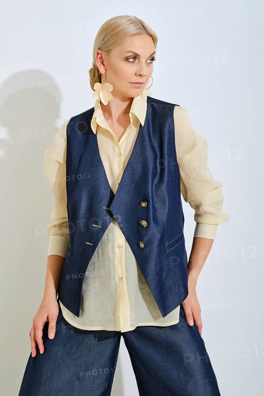 A fashion model poses confidently in a navy vest and light blouse. The background is bright and minimalist, emphasizing the trendy outfit in a casual yet fashionable way