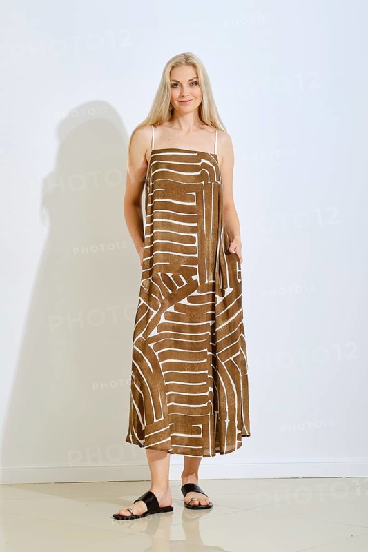 A woman stands confidently in a brown sundress featuring a unique white pattern. The setting is a bright, modern studio with minimal decor, highlighting her fashion choice and relaxed pose