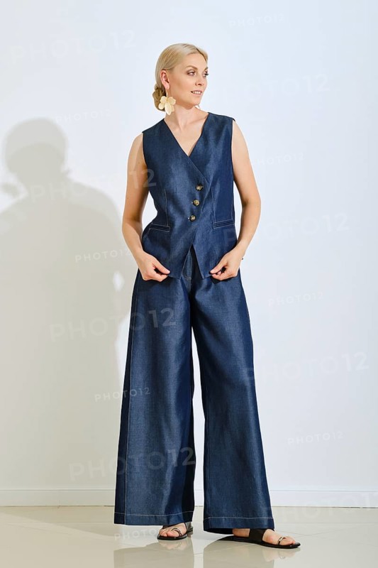 A woman stands gracefully in a stylish blue denim vest and wide-legged pants. She is indoors, showcasing her trendy outfit with a neutral background, highlighting her elegance and fashion sense