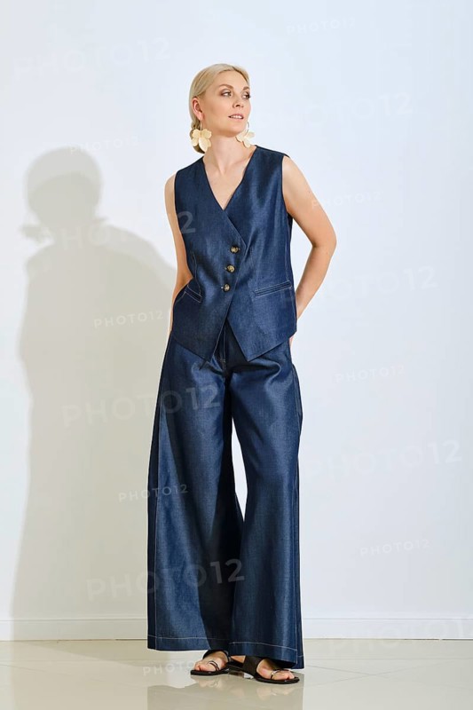 A fashionable woman stands confidently in a sleek denim vest and wide-leg pants. Her accessories complement the look, adding elegance. The setting is simple with a light background