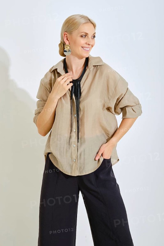 A woman is standing with a smile, dressed in a light beige shirt and dark pants. She wears a statement necklace and displays a relaxed yet fashionable look in a bright, minimalistic setting