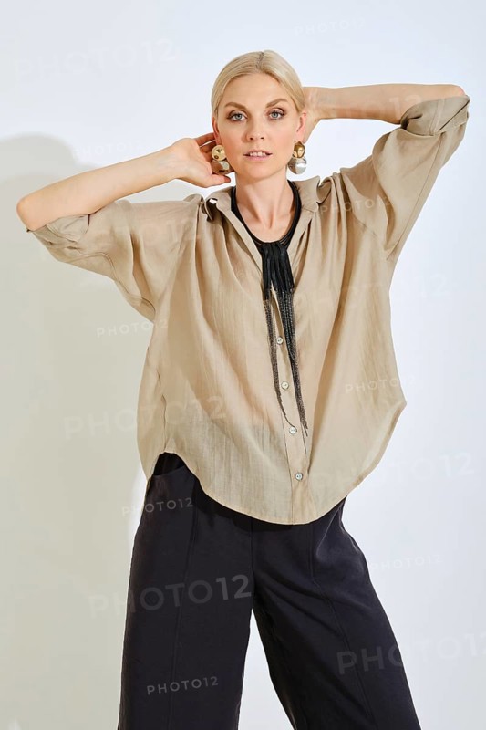 A woman strikes a confident pose while wearing a chic beige shirt complemented by bold accessories. The minimalist backdrop highlights her outfit, showcasing modern fashion aesthetics