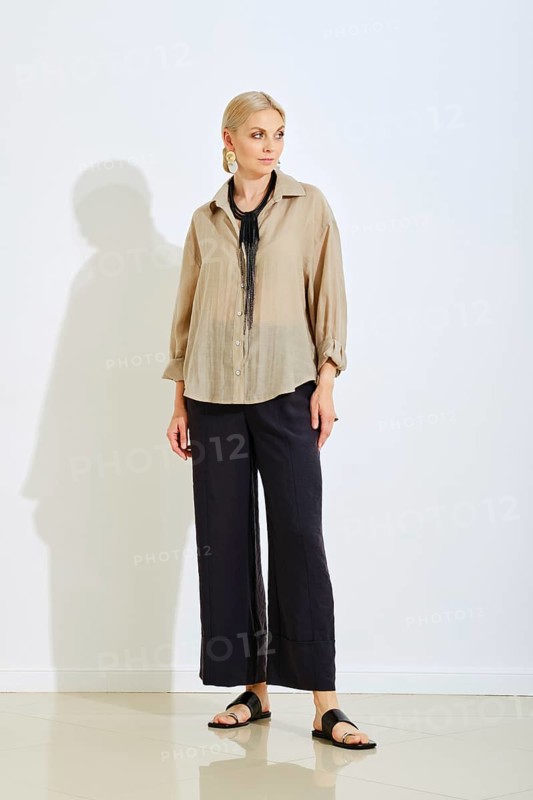 A model stands confidently in a bright studio space wearing a stylish beige top and black wide-leg pants. The hard light highlights her fashionable look, creating a relaxed yet sophisticated vibe
