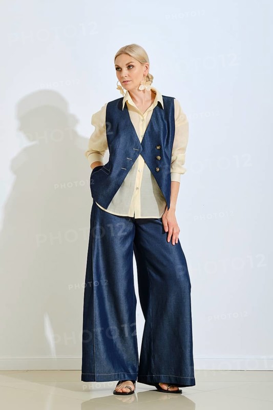 A fashion model stands confidently in a minimalist studio, wearing a denim vest and wide-leg pants paired with a light blouse. The soft lighting highlights her stylish look and elegant posture