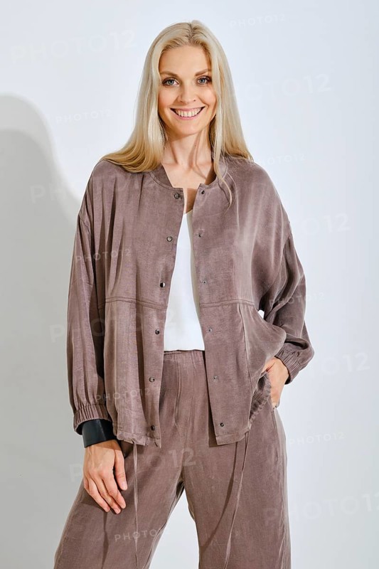 A smiling woman with long blonde hair is posing confidently in a soft brown casual outfit. She is wearing a light top and matching loose-fitting pants, standing against a plain light backdrop