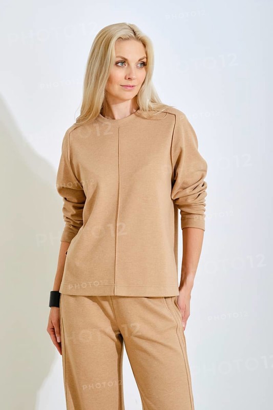 A confident woman stands elegantly in a bright studio, wearing a simple beige top and matching pants