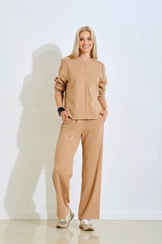 A cheerful woman is posing indoors wearing a light beige two-piece outfit that includes a loose-fitting top and wide-leg pants. Her long blonde hair and confident smile add charm to the scene