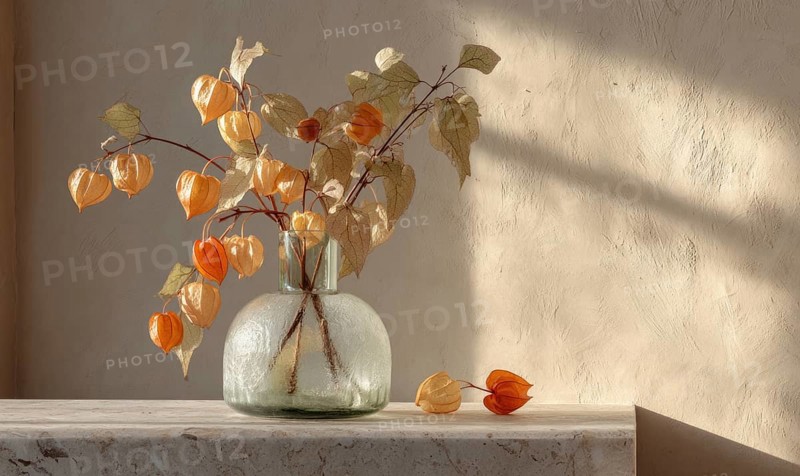 A vase of flowers sits on a table, with a few leaves scattered around it. The vase is filled with orange flowers, and the leaves are yellow. AI generated