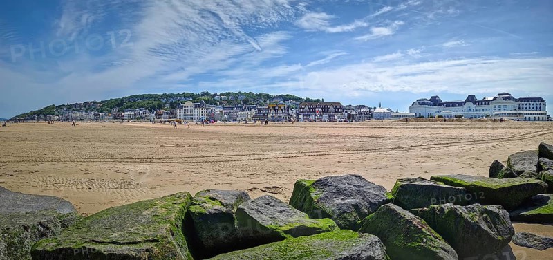 Panoramic view of Trouville-sur-Mer sandy beach, Normandy, France. Large, moss-covered breakwater rocks in foreground and charming town with traditional buildings in the background, Trouville-sur-Mer, France