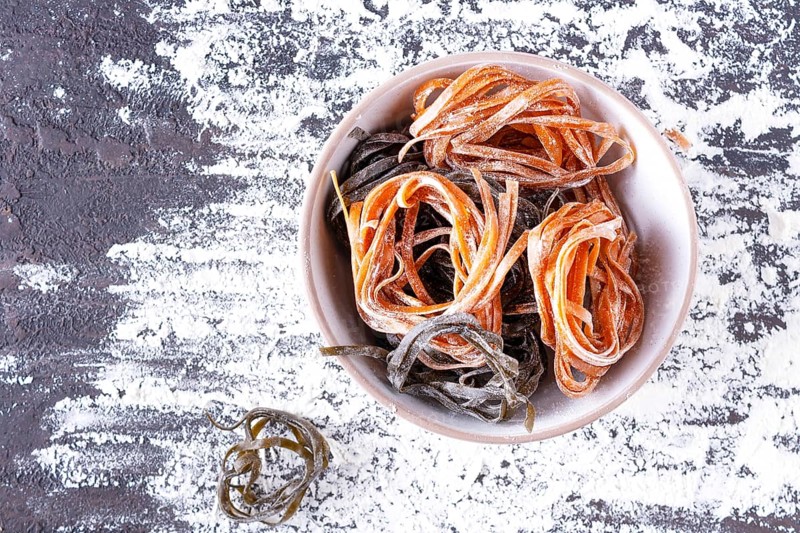 Pasta with carrots and spinach in a round plate on a table covered with flour, top view, space for text