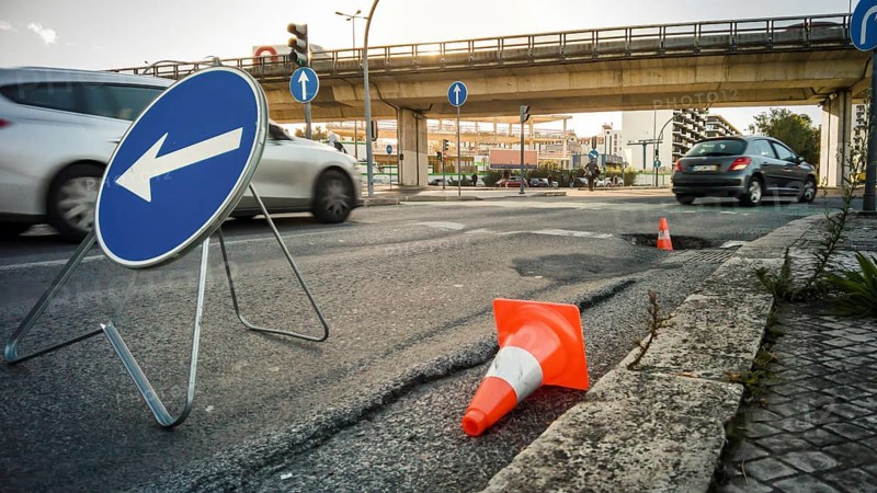City street with large pothole marked by traffic cones and detour sign, cars passing by and overpass in background. Urban infrastructure issues, road safety, busy traffic hours, street repairs, traffic disruption, heatwave, asphalt melting, summer, Lisbon, Portugal