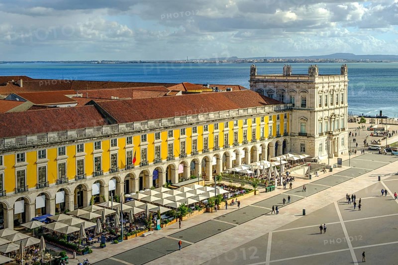 Scenic view of Commerce Square or Praca do Comercio in Lisbon, Portugal, iconic yellow Pombaline architecture, bustling outdoor cafes, Tagus River, city life, historic downtown, sunny day, sky with clouds. Sun ray over building, travel destination