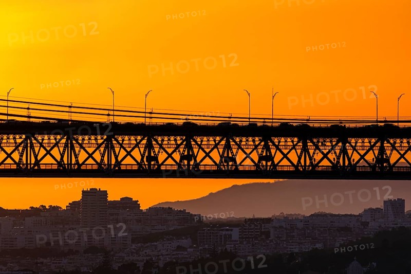 Bridge spans city skyline, silhouetted against vivid orange sunset. Soft city outlines and distant hills frame flowing late evening traffic, dynamic urban energy, tranquil evening light for unique harmony of architecture and nature. Urban landscape, Lisbon, Portugal