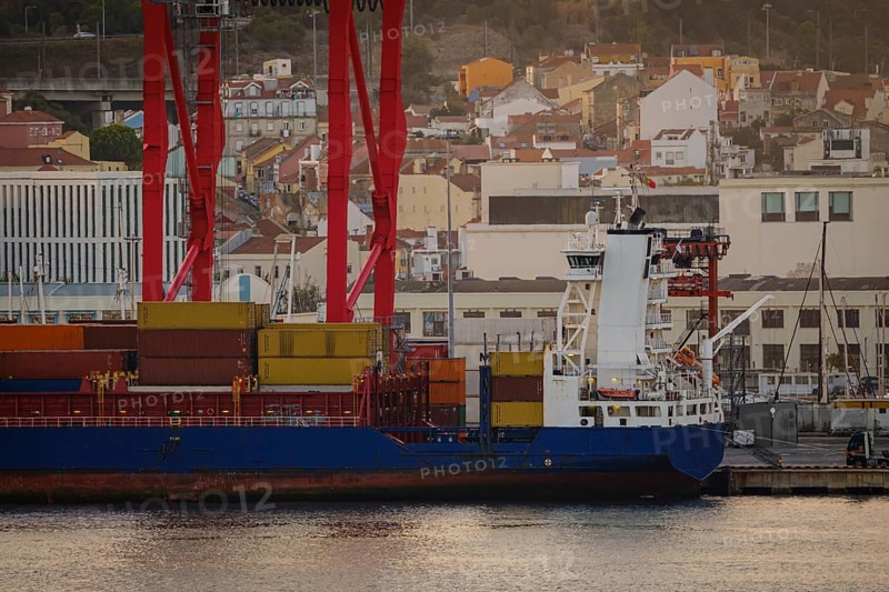 Cargo ship loaded with multicolored containers docked at Lisbon port, red cranes and city vibrant buildings on sunny evening. Dynamic residential and industrial scene by waterfront, urban skyline, loading, unloading, calm waters, commerce, industry