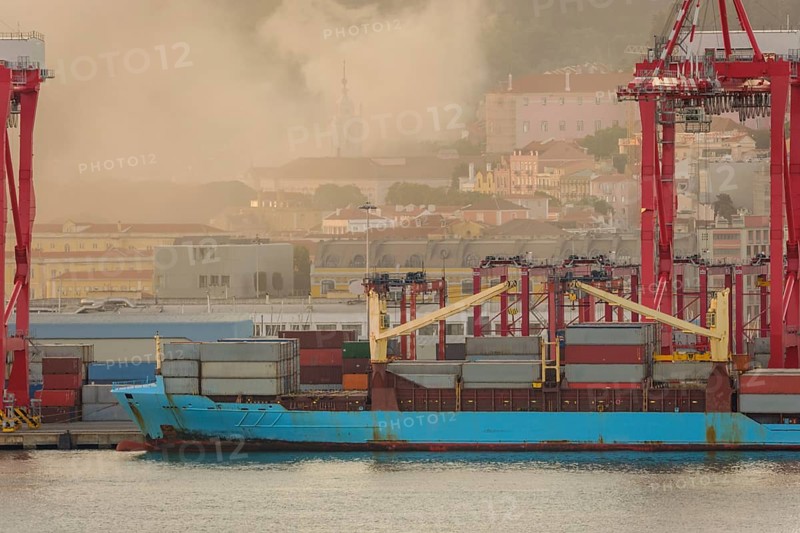 Blue container ship docked at Lisbon port, red cranes and stacks of shipping containers on deck, city buildings and church spire in evening haze and fire smoke in background. Risk of fires in summer, drought and heatwave, danger, damage, Portugal