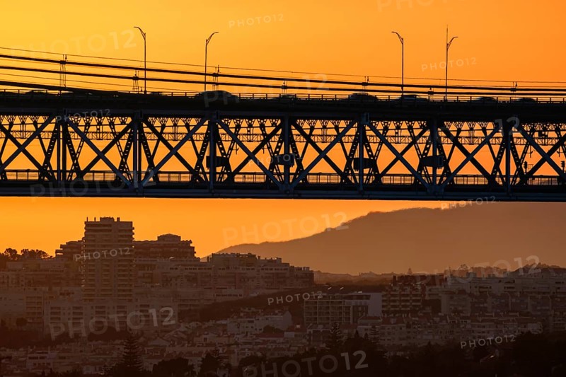 Striking close-up of famous 25 de Abril bridge, its silhouette against bright orange sunset sky. Lisbon, Portugal, cityscape and distant hills in hazy summer evening, blurred vehicles cross bridge, modern buildings, industrial structure, urban life