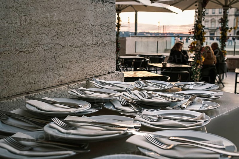 Plates neatly arranged with forks, knives, and napkins on stone countertop at outdoor restaurant in selective focus. Blurred background with European city square at sunset. Relaxed dining, people seated, umbrellas, travel destination, summer vacation, Lisbon, Portugal