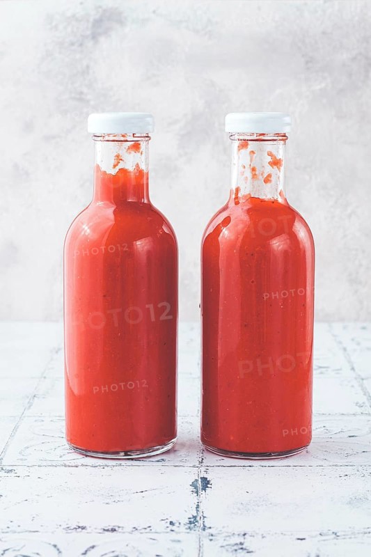 Two clear glass bottles hold vibrant red sauce, likely a spicy or tangy condiment, in a well-lit kitchen area. The smooth surface enhances the color and freshness of the sauce