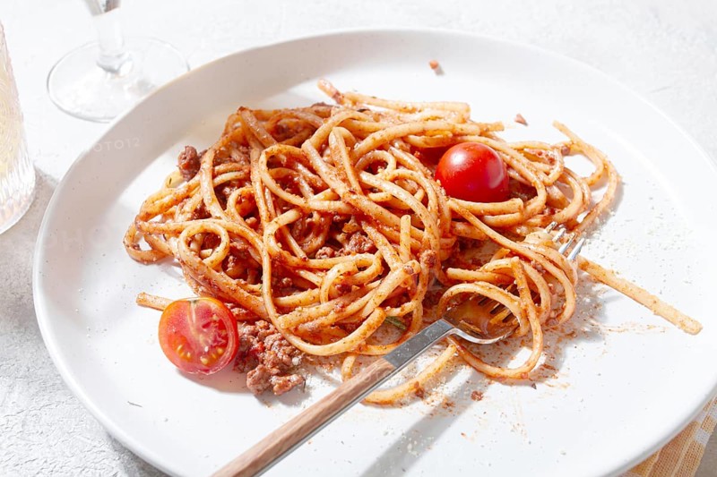 A plate holds freshly cooked spaghetti topped with flavorful meat sauce and garnished with bright cherry tomatoes. A glass sits nearby, reflecting a relaxed dining atmosphere