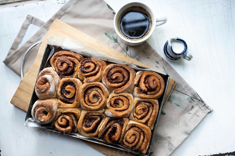 Freshly baked cinnamon rolls on a wooden board with a small pot of milk and a cup of coffee