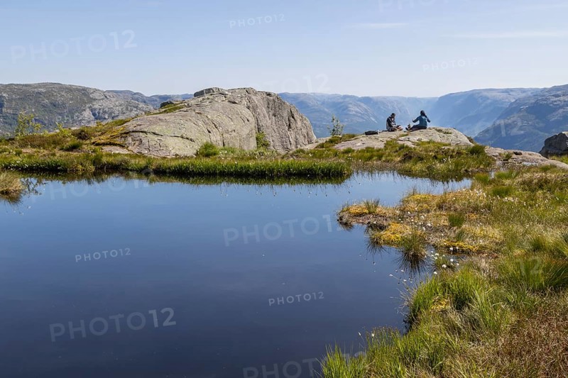 Two people sitting on a rock by a mountain lake, above the Preikestolen hiking trail and high above Lysefjord, beneath a blue summer sky and surrounded by the vast, barren landscape of the Scandinavian Mountains. Strand, Rogaland in Vestlandet, the Western Norway