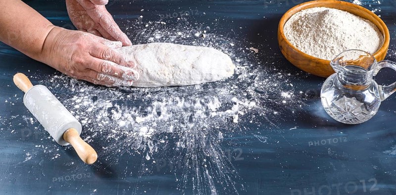 Female hands preparing fresh artisanal bread dough on a dark kitchen table. Baking ingredients flour, water, and a rolling pin are present