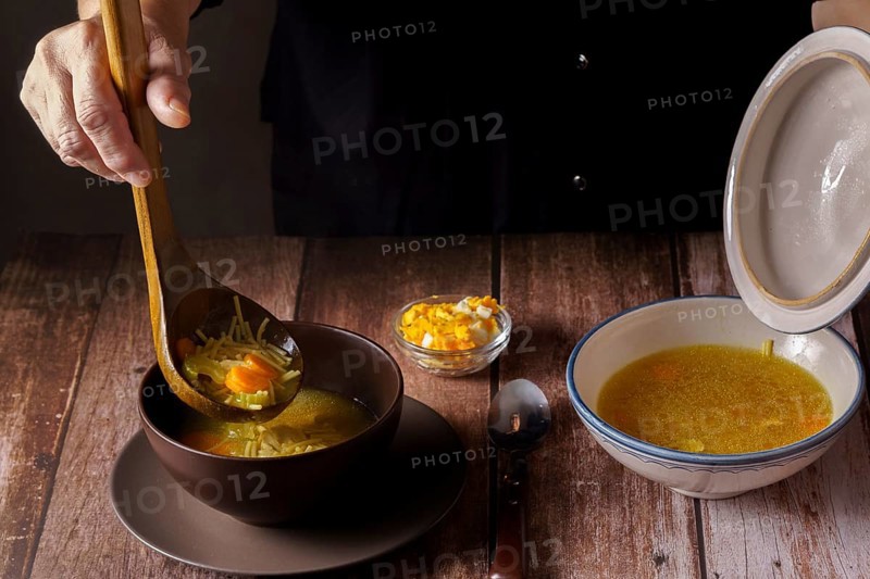 Woman serving chicken soup with a wooden spoon in a brown bowl on a wooden table