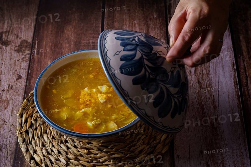Woman opening the lid of a tureen with chicken noodle soup and vegetables on a wooden table