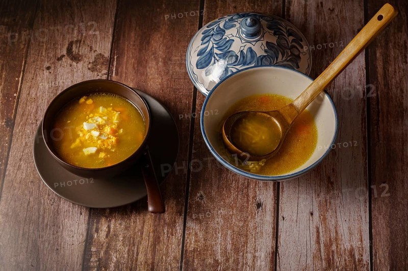 Chicken noodle soup in a white soup tureen and a brown bowl with a plate and spoon on a wooden table