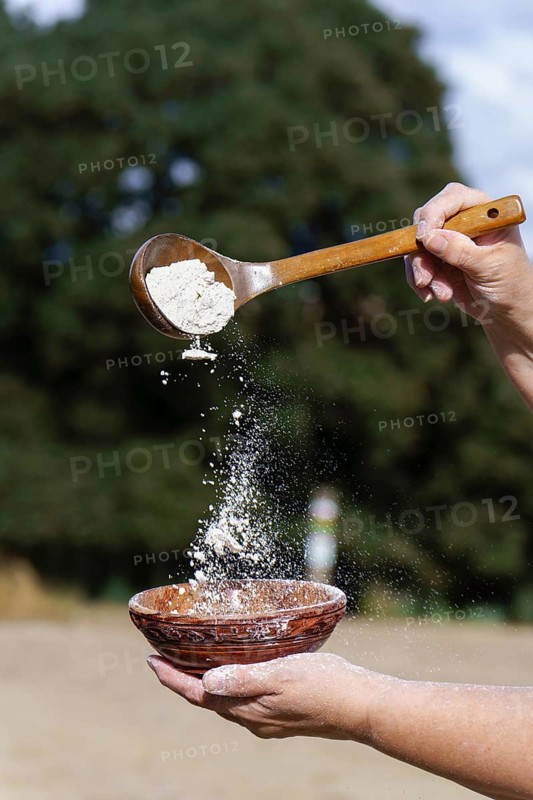 Hands sifting white flour from a wooden spoon into a rustic ceramic bowl, preparing ingredients for baking
