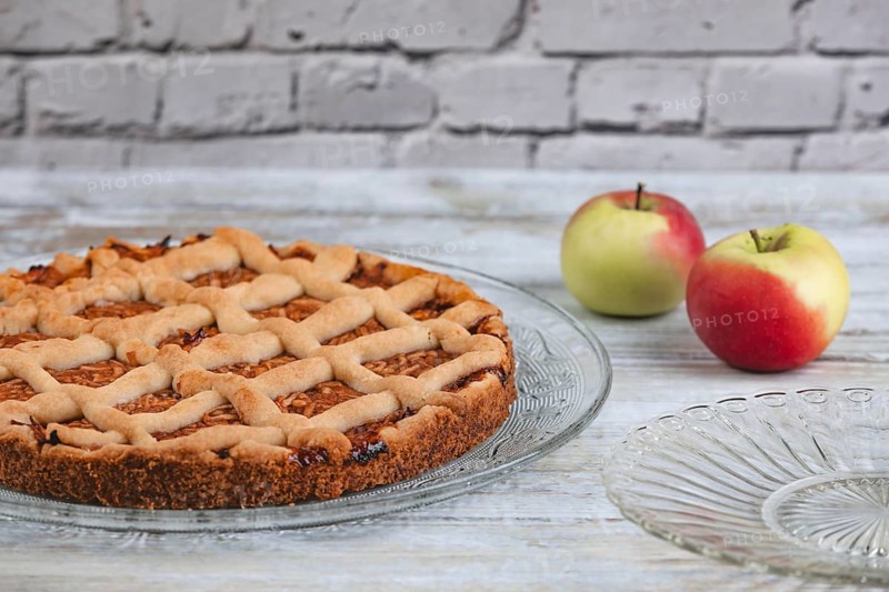 Apple cake on a glass plate with two apples next to it in front of a rustic brick wall