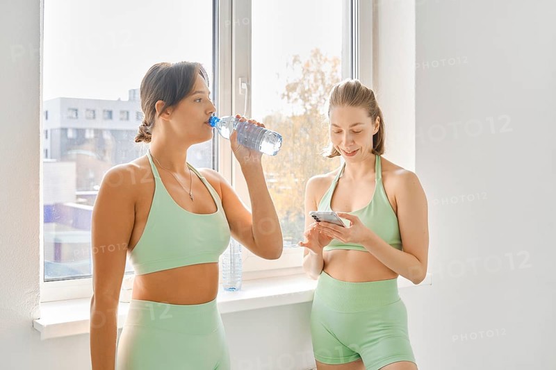 Two women take a break during a fitness stretching class in a bright indoor studio. One drinks water while the other checks her phone, enjoying the pause in their workout session