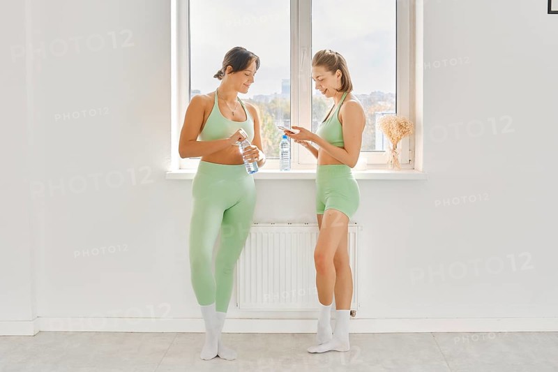 Two women pause during their fitness stretching class in a bright and spacious studio. They are dressed in matching light green workout outfits and relax by chatting and checking their phones