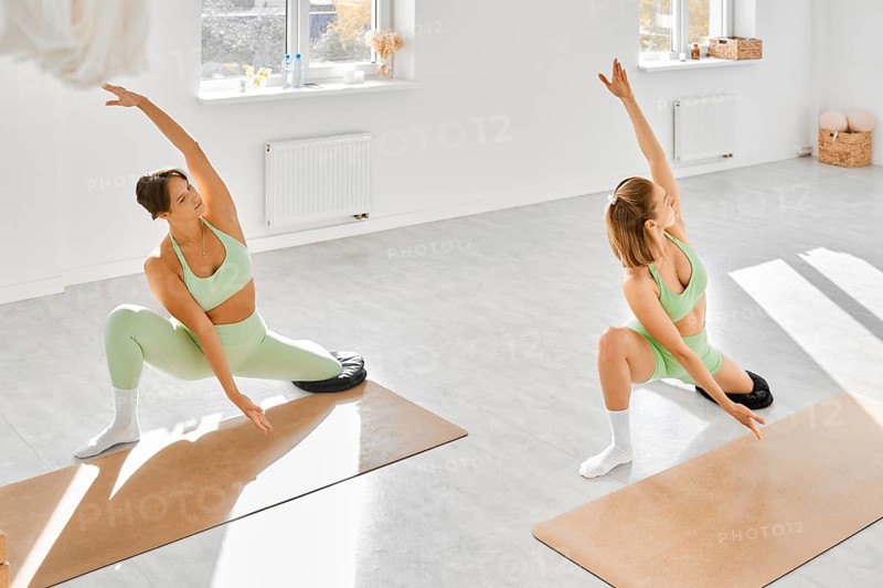 Two women are engaged in a yoga session in a well-lit studio filled with natural light. They perform flexible poses on mats while wearing matching green outfits. The atmosphere is calm and focused