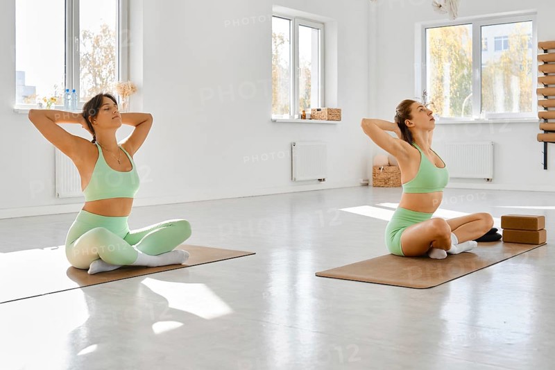 Two women are seated on yoga mats in a spacious, well-lit studio. They are dressed in matching light green outfits, focusing on stretching exercises. The serene environment promotes relaxation