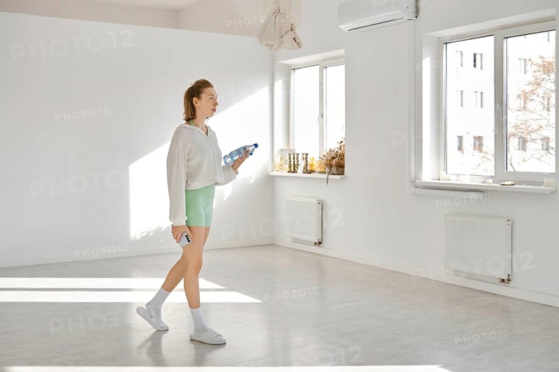 In a bright studio with white walls and sunlight streaming through large windows, a young woman walks with drinking water bottles