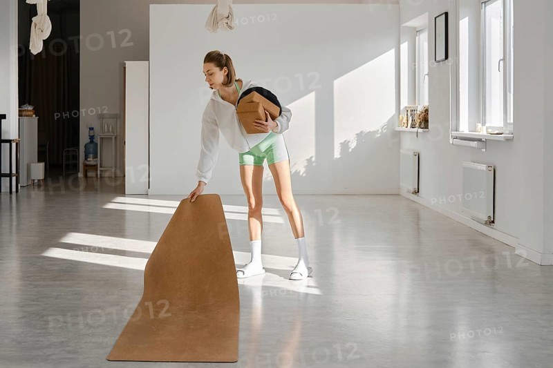 A young woman in light clothing is setting up a yoga mat in a spacious, sunlit studio. She is carrying a box and appears focused on starting her workout routine