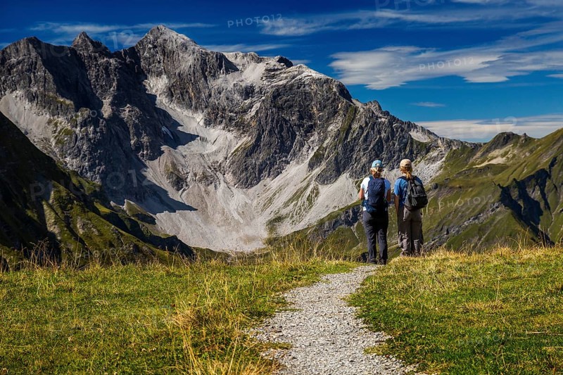 Two hikers stand in front of an impressive mountain backdrop under clear skies, Schröcken, Austria