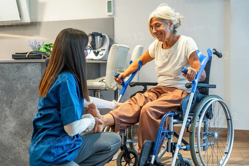 Nursing professional administering physical therapy to an elderly woman in a wheelchair, focusing on leg rehabilitation and providing home care support for recovery