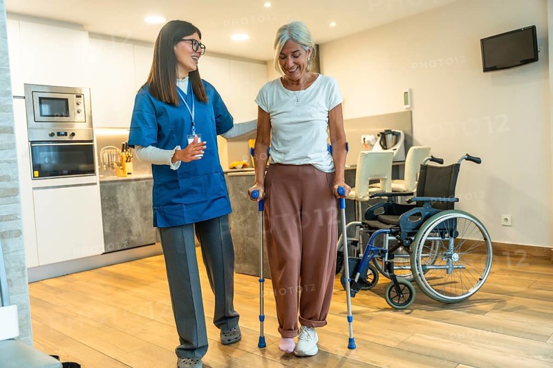 Young female nurse helping a happy elderly woman recovering from injury, walking with crutches during home care physiotherapy for rehabilitation and independent living
