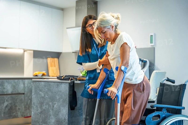 Young female caregiver is providing support and assistance to a senior woman, helping her during a home rehabilitation session, using a pair of crutches in a kitchen setting