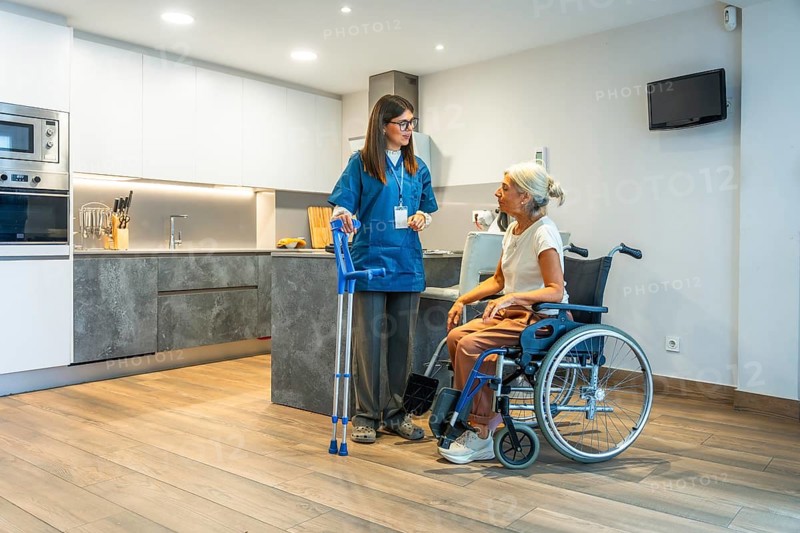 Professional caregiver providing home assistance to a senior woman in a wheelchair, discussing her health and well being in a modern kitchen with medical crutches nearby
