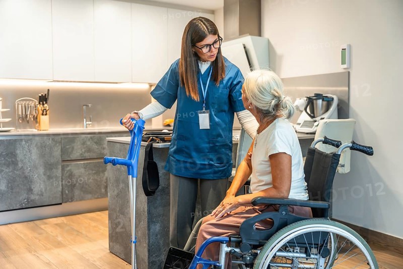 Young female caregiver in scrubs assisting a senior woman in a wheelchair, providing home healthcare support and discussing mobility with crutches in a modern kitchen setting