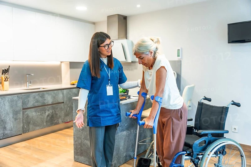 Home nurse assisting a senior woman using crutches to walk, providing support and physical therapy during her rehabilitation process in a modern home environment