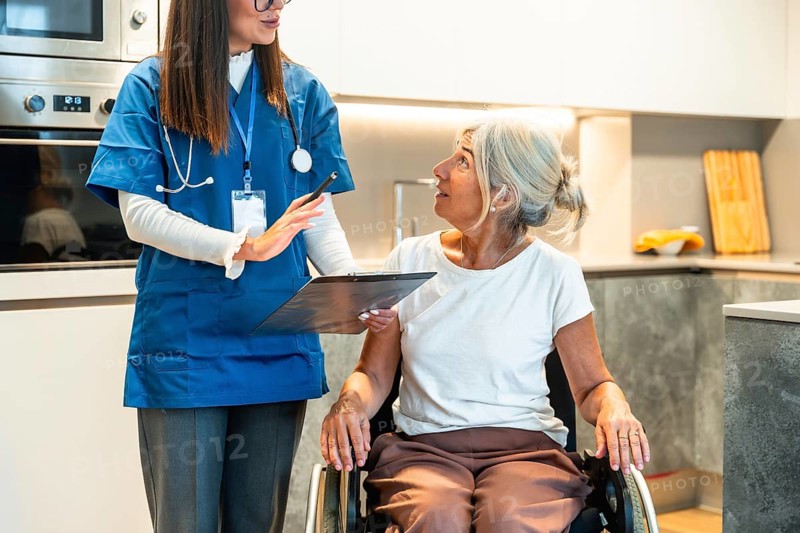 Nurse reviewing a healthcare plan with an elderly woman in a wheelchair in a modern kitchen, providing compassionate home care, support and guidance for independent living