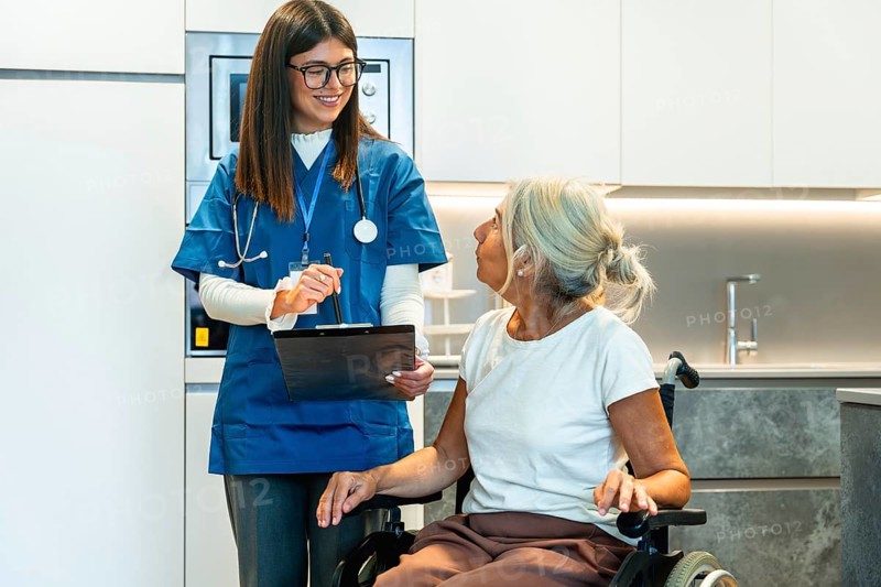 Young nurse wearing a stethoscope and holding a clipboard, discussing a treatment plan with a smiling senior woman sitting in a wheelchair for home care