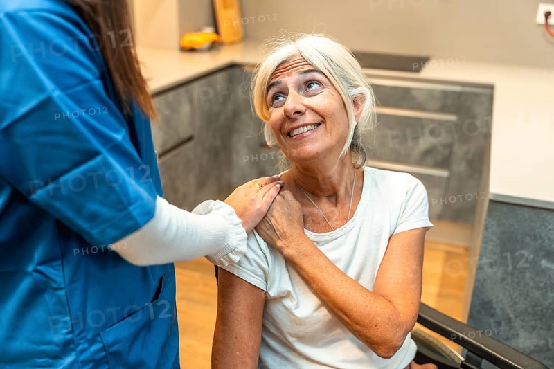 Healthcare professional in scrubs placing a comforting hand on the shoulder of a smiling senior woman with grey hair, illustrating elderly support and compassionate care in a home setting
