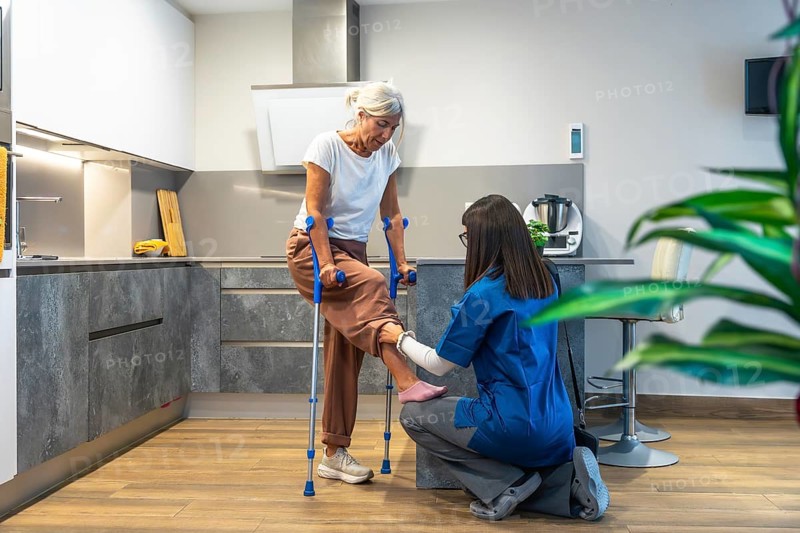 Healthcare professional guiding elderly woman with crutches during home physiotherapy session, supporting mobility, recovery and independence in a comfortable domestic setting