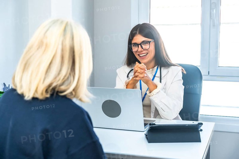Young female doctor wearing a lab coat and stethoscope, smiling while actively listening to a patient during a healthcare consultation in a bright clinic office